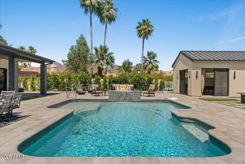 View from the Scottsdale home's kitchen toward the resort-style backyard, where a sparkling pool with Baja shelf, spacious paver decking, multiple seating areas, mature landscaping, and the separate guest house create an inviting setting for outdoor living and entertaining.