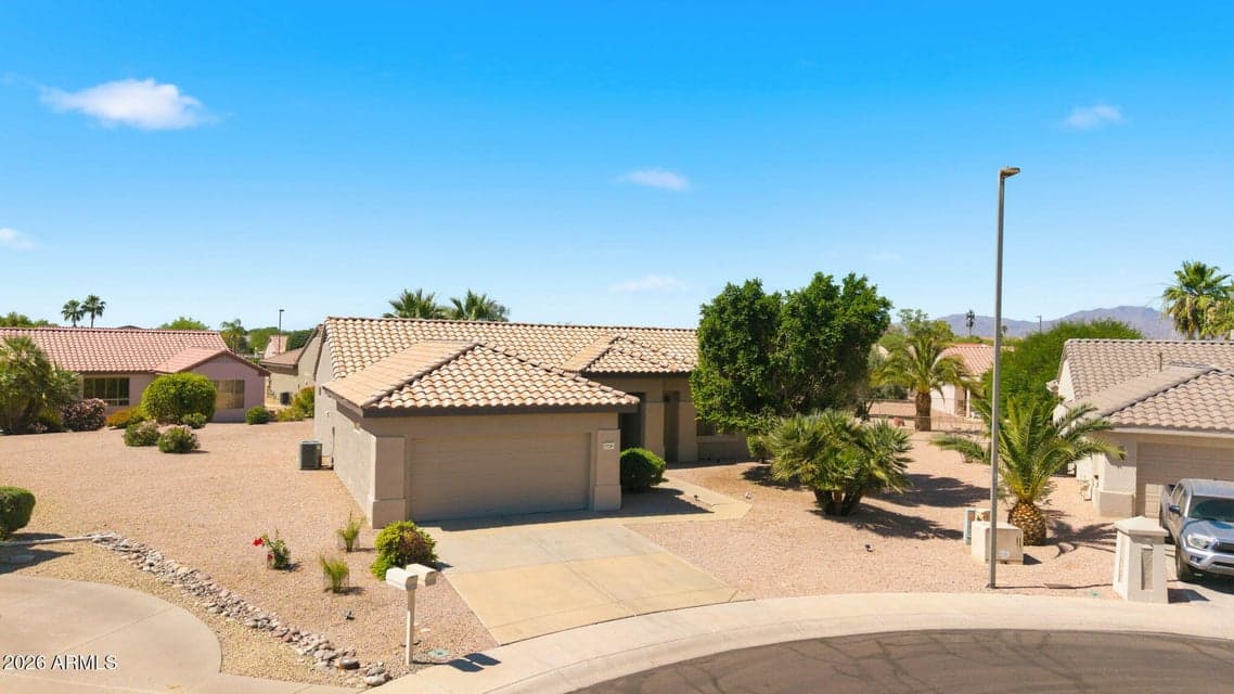 Aerial front view of this Surprise property showcasing its single-story design, tile roof, attached garage, spacious driveway, mature palm trees, and low-maintenance desert landscaping. The setting offers classic Arizona curb appeal with an inviting approach to the home.