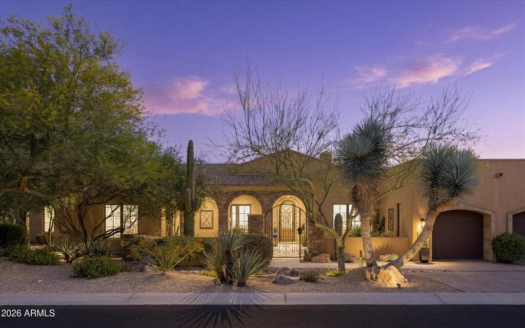 A striking twilight exterior showcasing the home’s desert setting, arched entry details, and inviting courtyard approach.