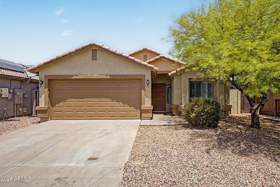 Inviting front exterior of this Tolleson home featuring a two-car garage, tile roof, mature shade tree, desert landscaping, and a welcoming covered entry.