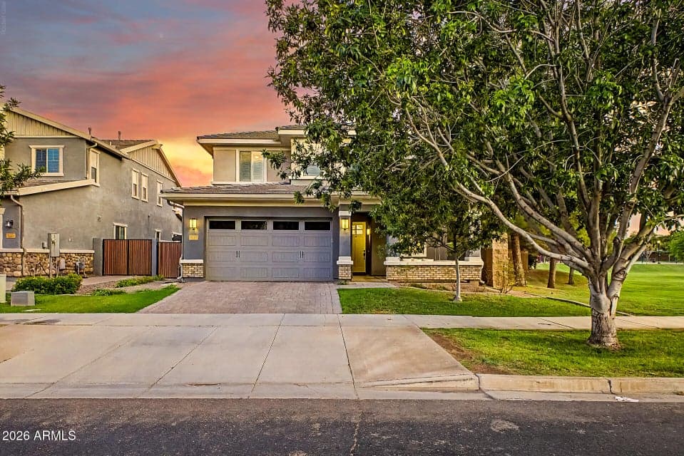 Exterior front, dusk — Two-story home with painted stucco exterior, gray carriage-style garage door, paver driveway, and yellow front door. Mature shade tree and adjacent green space visible.