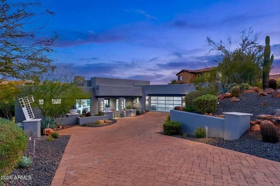 Striking contemporary curb appeal featuring a paver driveway, translucent glass garage door, and mature low-maintenance desert landscaping illuminated by dramatic evening lighting.