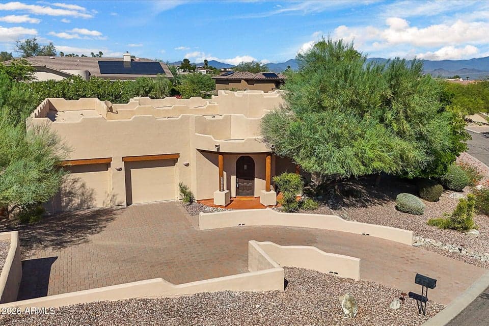 Welcoming classic Pueblo-style architecture, paver driveway, low-maintenance desert xeriscaping, and beautiful mountain backdrop under bright, Arizona skies.