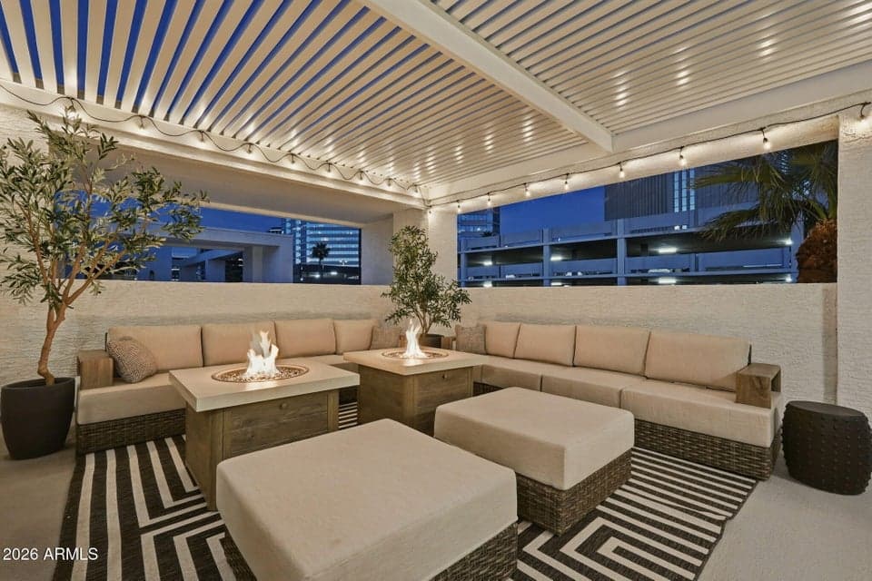 Twin gas fire pit tables anchor the rooftop lounge, framed by a louvered pergola with string lighting overhead and a black-and-white striped indoor-outdoor rug underfoot. Deep wicker-base sectional seating and olive trees in planters wrap the space, with downtown Phoenix high-rises and a parking structure visible beyond the stucco parapet at dusk. Part of the private rooftop level at 128 W Willo Ct, Phoenix, AZ 85003.